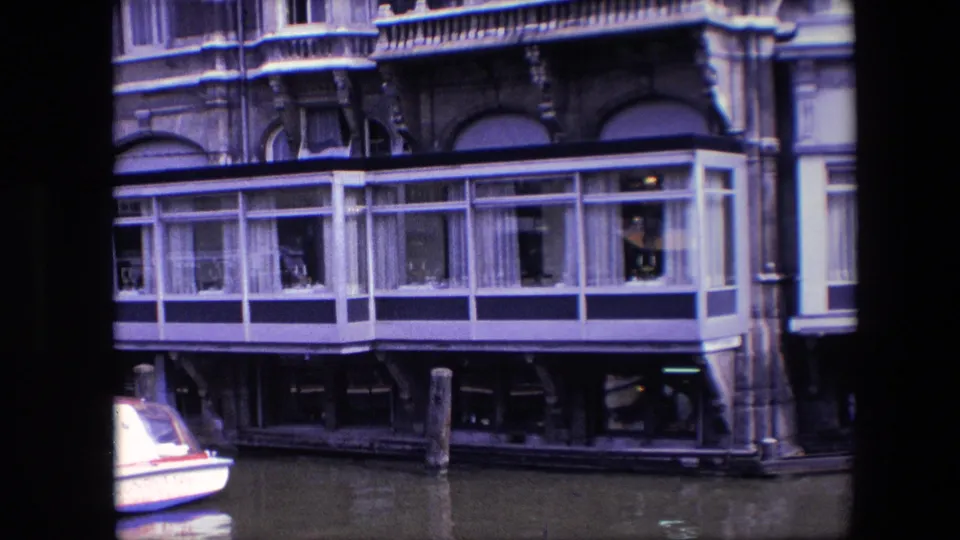 1967 Amsterdam home-movie frame showing a canal houseboat with a glass-enclosed room, white curtains, dark lower supports, mooring posts, and a partial white boat at left.