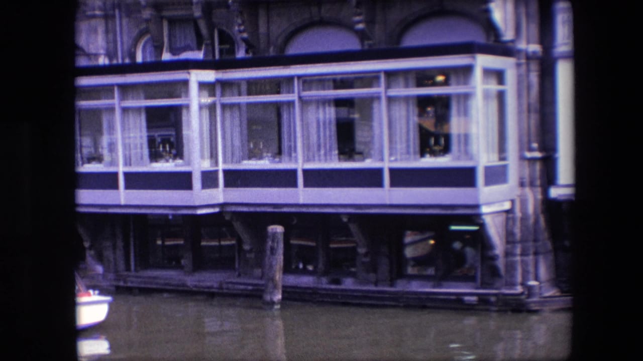 1967 Amsterdam canal view of a houseboat's glass room with white framing, curtains, small interior objects, dark supports, and a wooden mooring post in front.