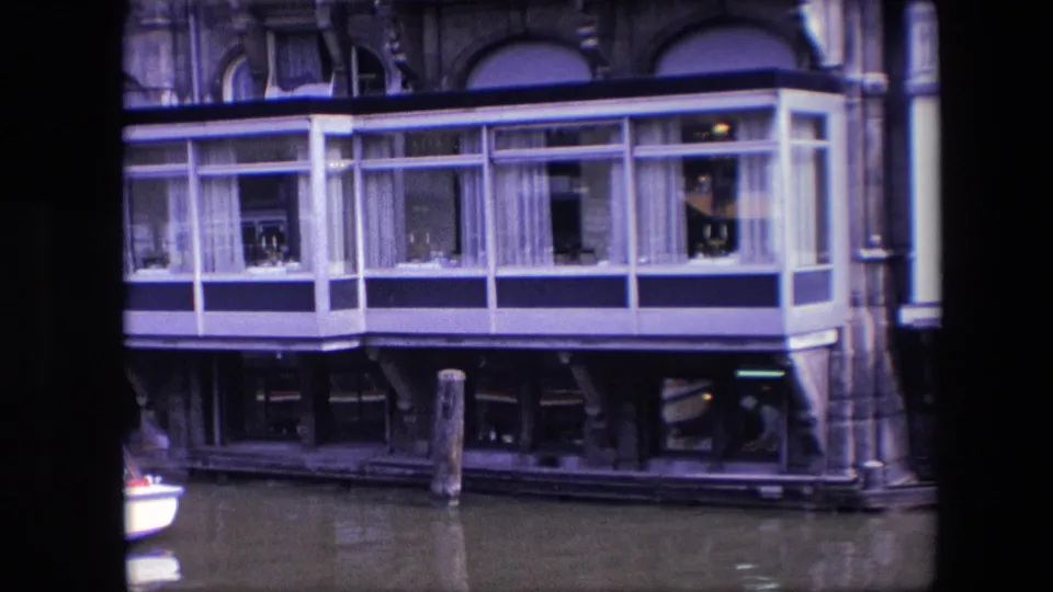 1967 Amsterdam canal view of a houseboat's glass room with white framing, curtains, small interior objects, dark supports, and a wooden mooring post in front.
