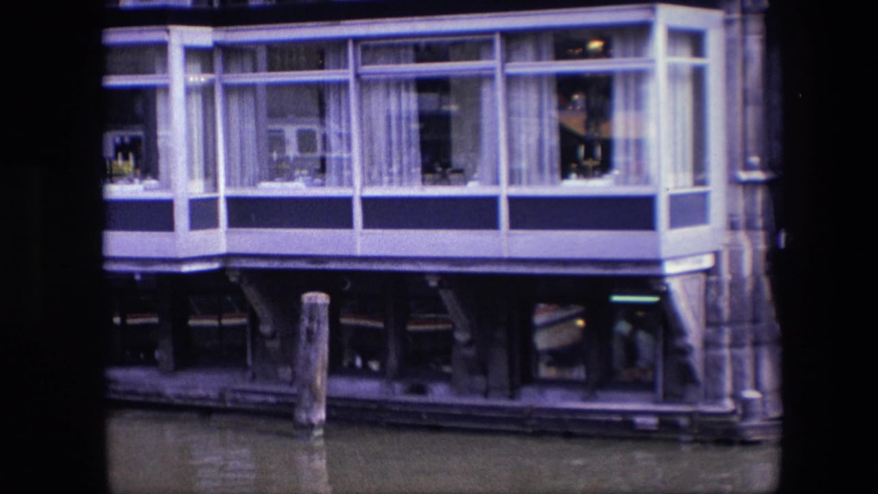 Closer 1967 Amsterdam frame of the houseboat's windowed living space, with white curtains, glass reflections, dark structural supports, and canal water below.