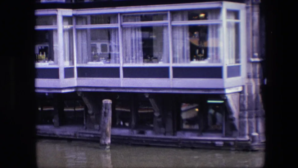 Closer 1967 Amsterdam frame of the houseboat's windowed living space, with white curtains, glass reflections, dark structural supports, and canal water below.