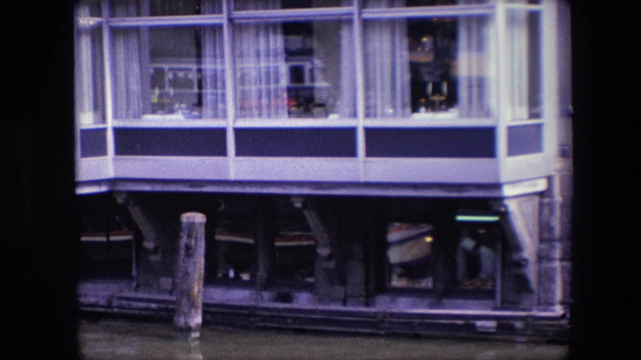 1967 Amsterdam home-movie frame showing the houseboat facade from across the canal, including broad windows, curtains, possible interior occupants or objects, a mooring post, and dark lower framing.