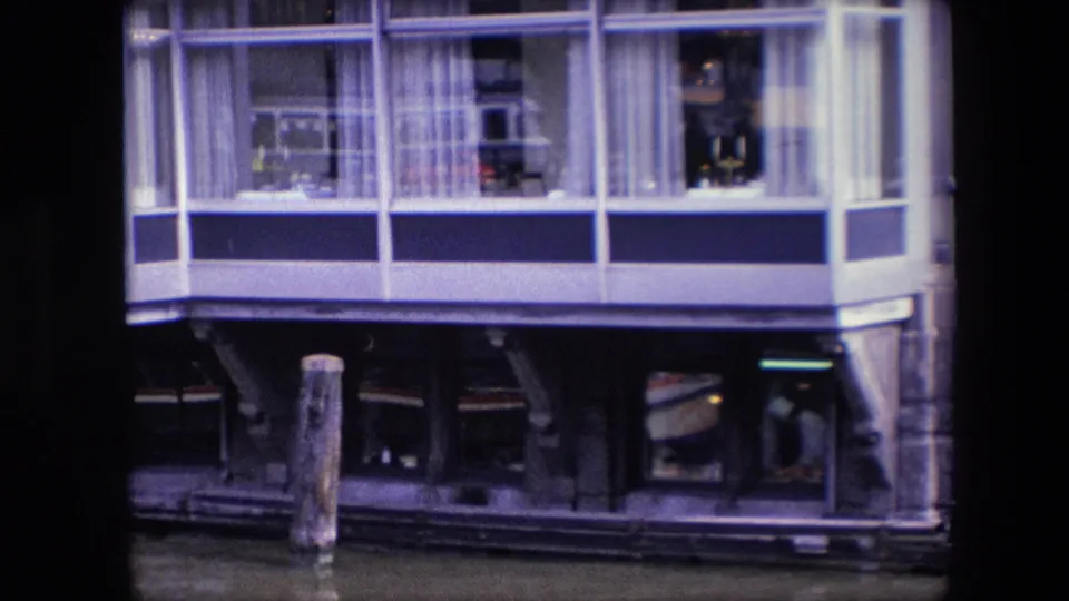 1967 Amsterdam home-movie frame showing the houseboat facade from across the canal, including broad windows, curtains, possible interior occupants or objects, a mooring post, and dark lower framing.