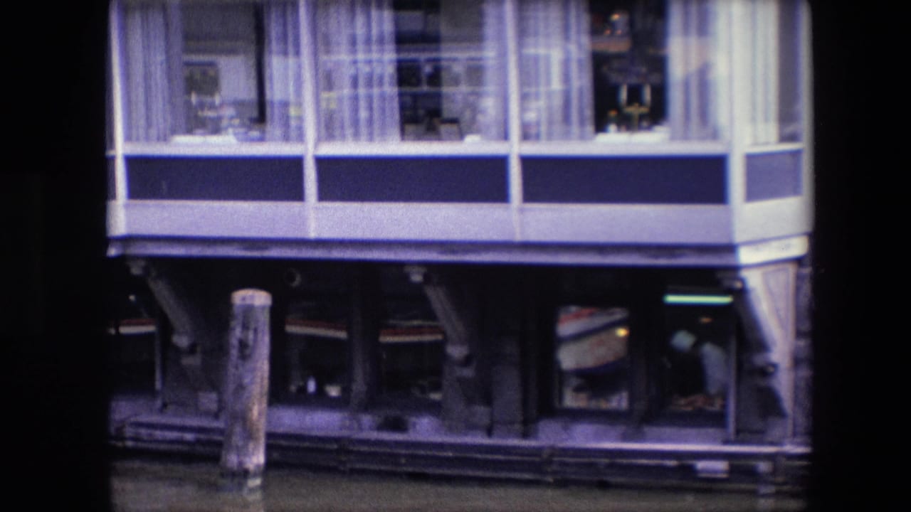 1967 archival home-movie frame of a blue-and-white waterfront building, likely in Amsterdam per metadata, with upper windows, curtains, dark lower openings, and a wooden piling in the water.