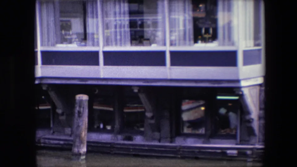 1967 archival home-movie frame of a blue-and-white waterfront building, likely in Amsterdam per metadata, with upper windows, curtains, dark lower openings, and a wooden piling in the water.
