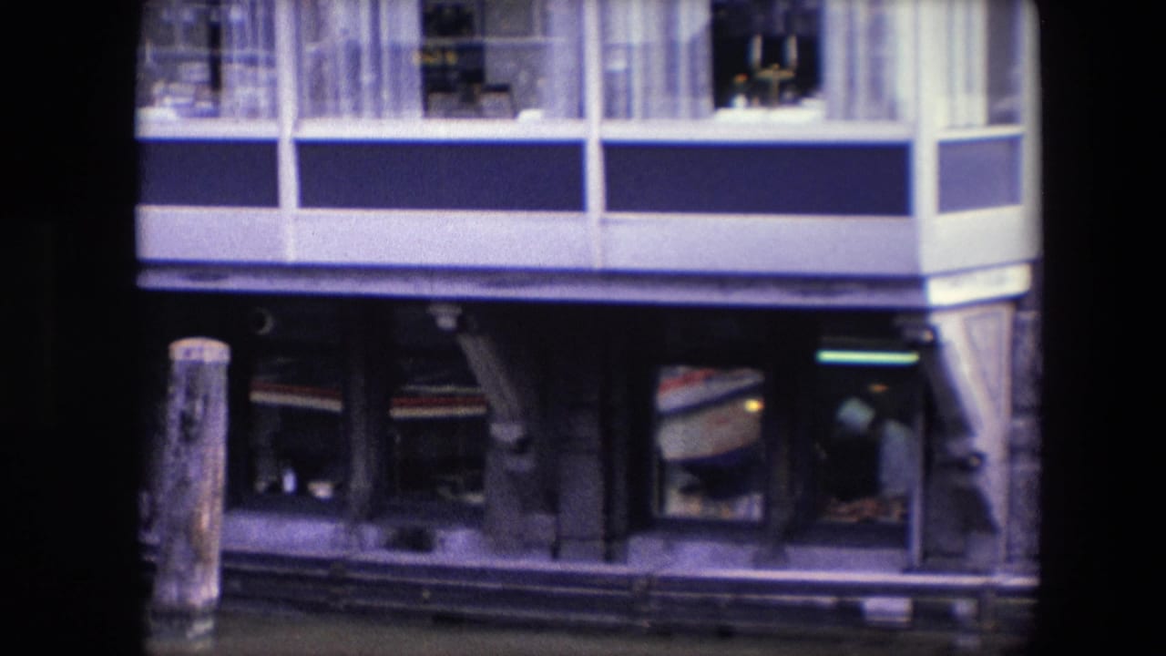Archival frame of the waterfront building during a slow zoom, showing the blue-and-white upper facade, lower restaurant windows, and a person visible inside near the right side.