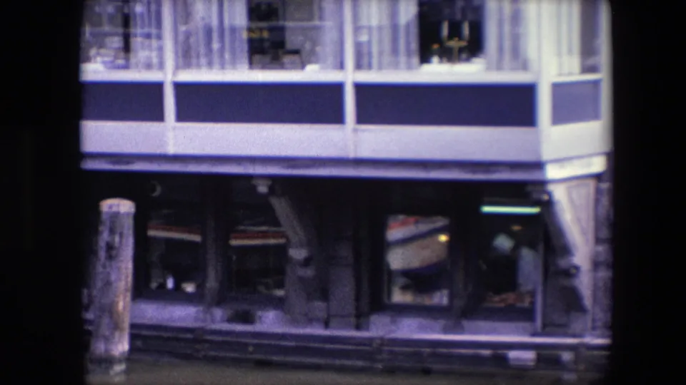 Archival frame of the waterfront building during a slow zoom, showing the blue-and-white upper facade, lower restaurant windows, and a person visible inside near the right side.