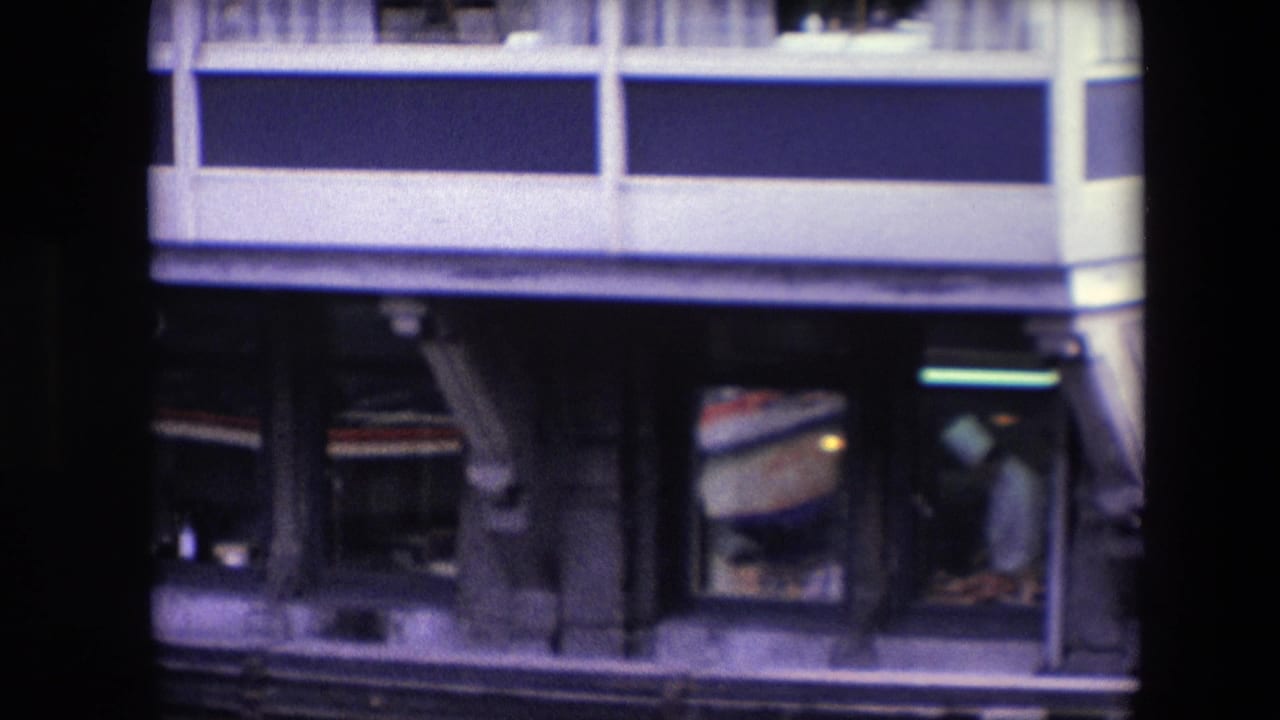 Closer 8mm frame of the lower waterfront facade, with a white-clad chef-like figure visible through a restaurant window and architectural supports above the waterline.
