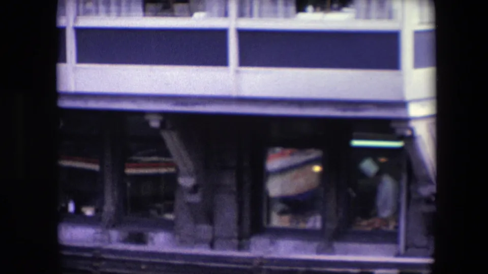Closer 8mm frame of the lower waterfront facade, with a white-clad chef-like figure visible through a restaurant window and architectural supports above the waterline.