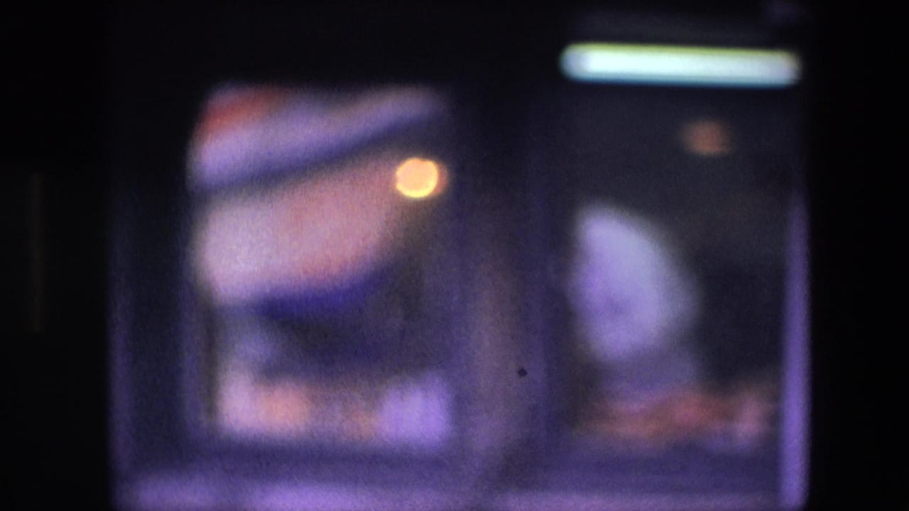 Blurred 8mm view of the restaurant interior, showing a light-shirted man near the right side and indistinct food or counter objects along the bottom edge.