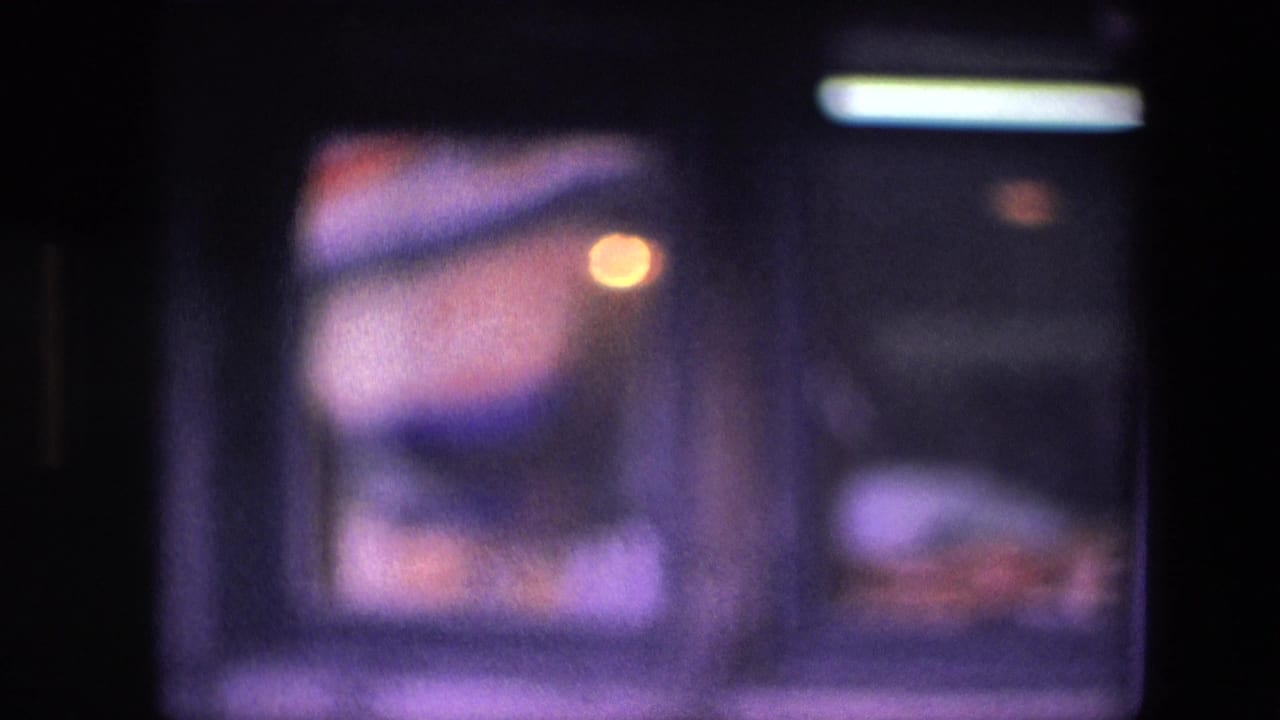 Final 1967 Amsterdam restaurant frame showing the worker lower in the window area, with dim interior light, dark framing, and soft purple-tinted film color.