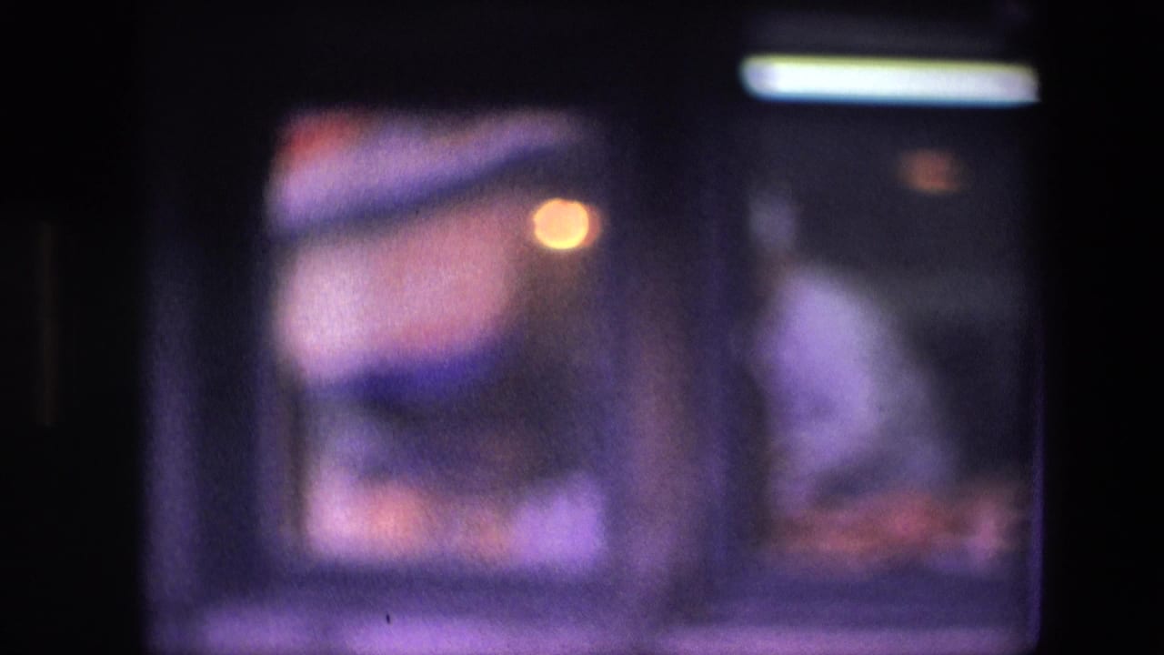 Softly focused 1960s 8mm frame of a dim restaurant kitchen window, with a cook in white partly visible behind reflective glass and cool overhead light.