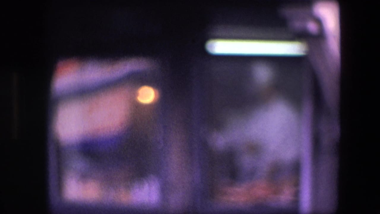 Archival home-movie frame showing a cook in white inside a dim kitchen, viewed through divided window glass with purple-blue low-light color.