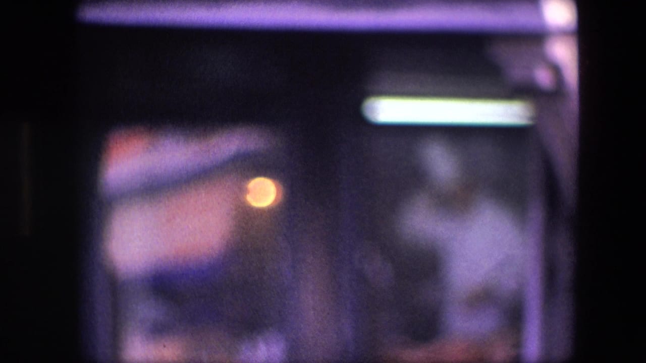 Low-light 8mm frame of a cook near a restaurant kitchen work area, with reflective glass, warm orange glow, and cool light above.