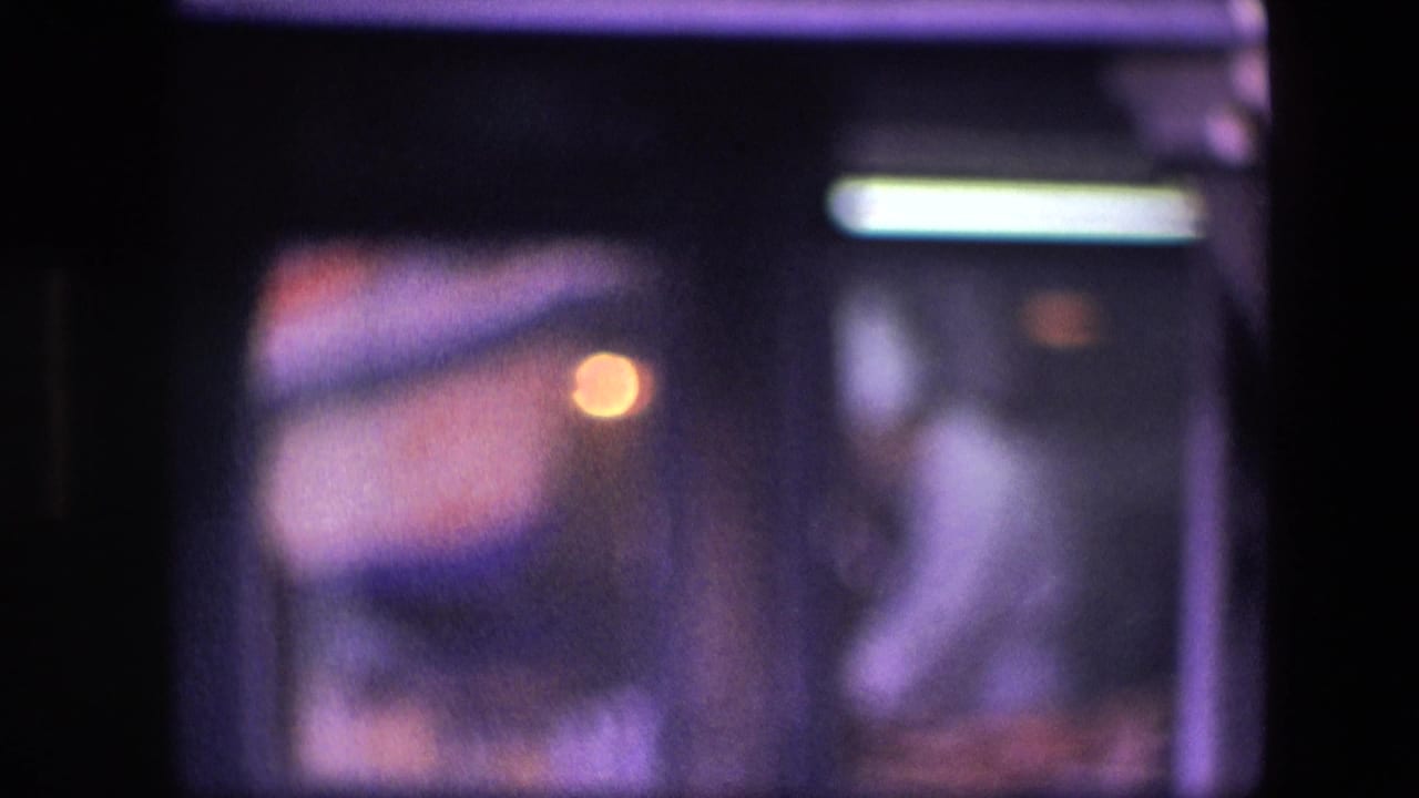 Soft, grainy 8mm frame of a cook in white behind a dark kitchen window, with an orange burner-like glow and cool fluorescent-style light.