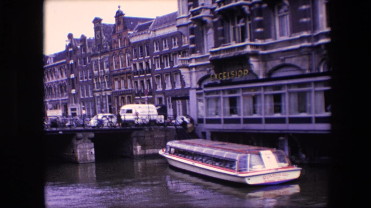 1967 8mm view of an Amsterdam canal with a white and red tour boat, stone bridge, parked vehicles, pedestrians, and canal-side buildings with an EXCELSIOR sign.