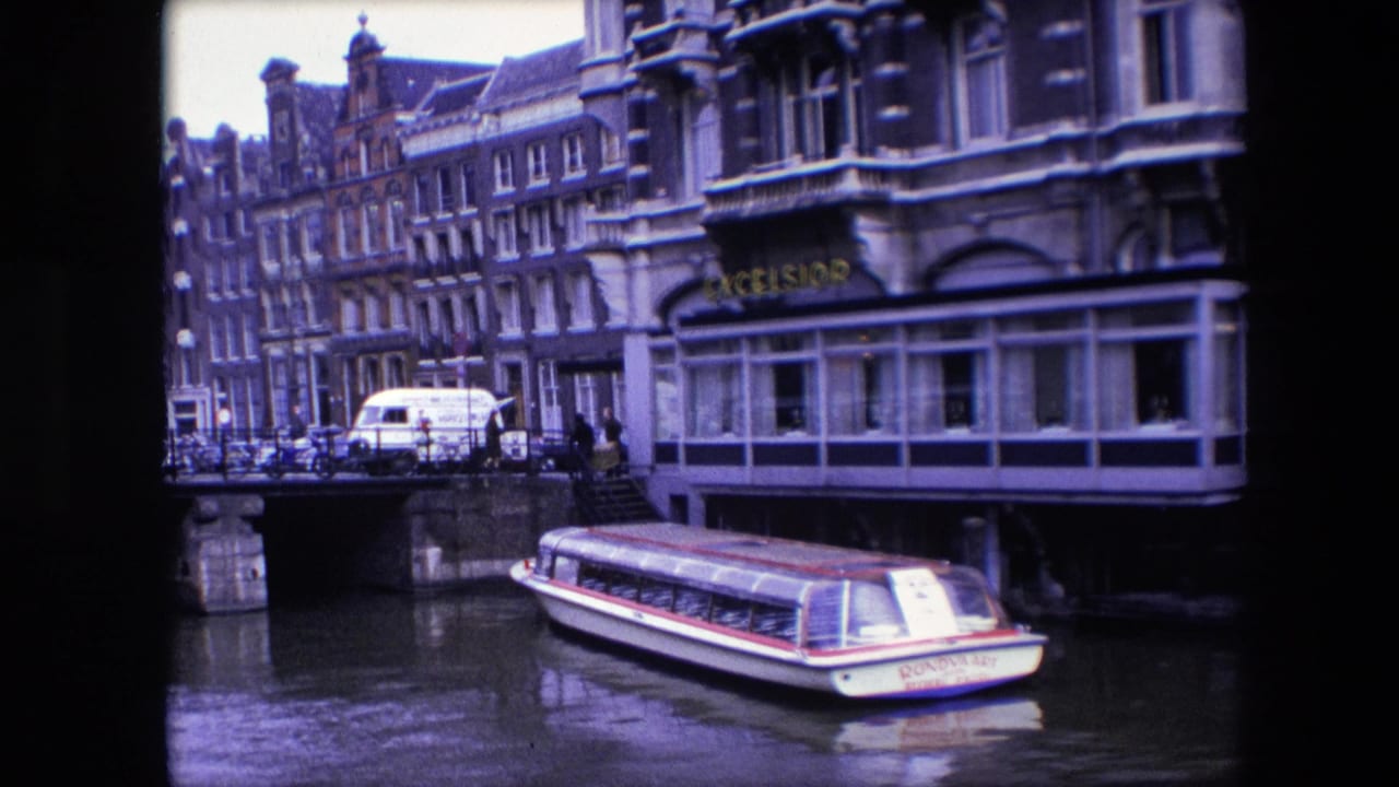 1967 Amsterdam canal footage with a tour boat in the foreground, a bridge at left, pedestrians near the railing, parked vehicles, and ornate building facades along the water.