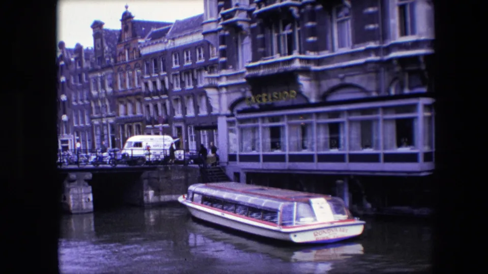 1967 Amsterdam canal footage with a tour boat in the foreground, a bridge at left, pedestrians near the railing, parked vehicles, and ornate building facades along the water.