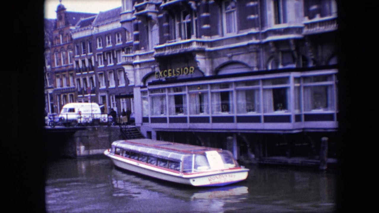 1967 Amsterdam travel footage frame showing a white rondvaart canal boat with red trim resting near the Excelsior hotel frontage and canal-side buildings.