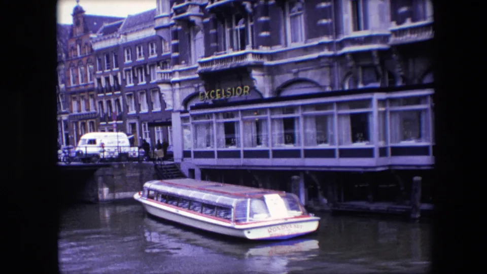 1967 Amsterdam travel footage frame showing a white rondvaart canal boat with red trim resting near the Excelsior hotel frontage and canal-side buildings.