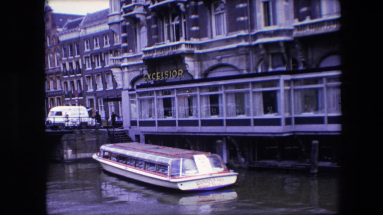 1967 Amsterdam canal view with a white rondvaart boat below the Excelsior hotel sign, glass-fronted windows, and canal-side street activity in the background.