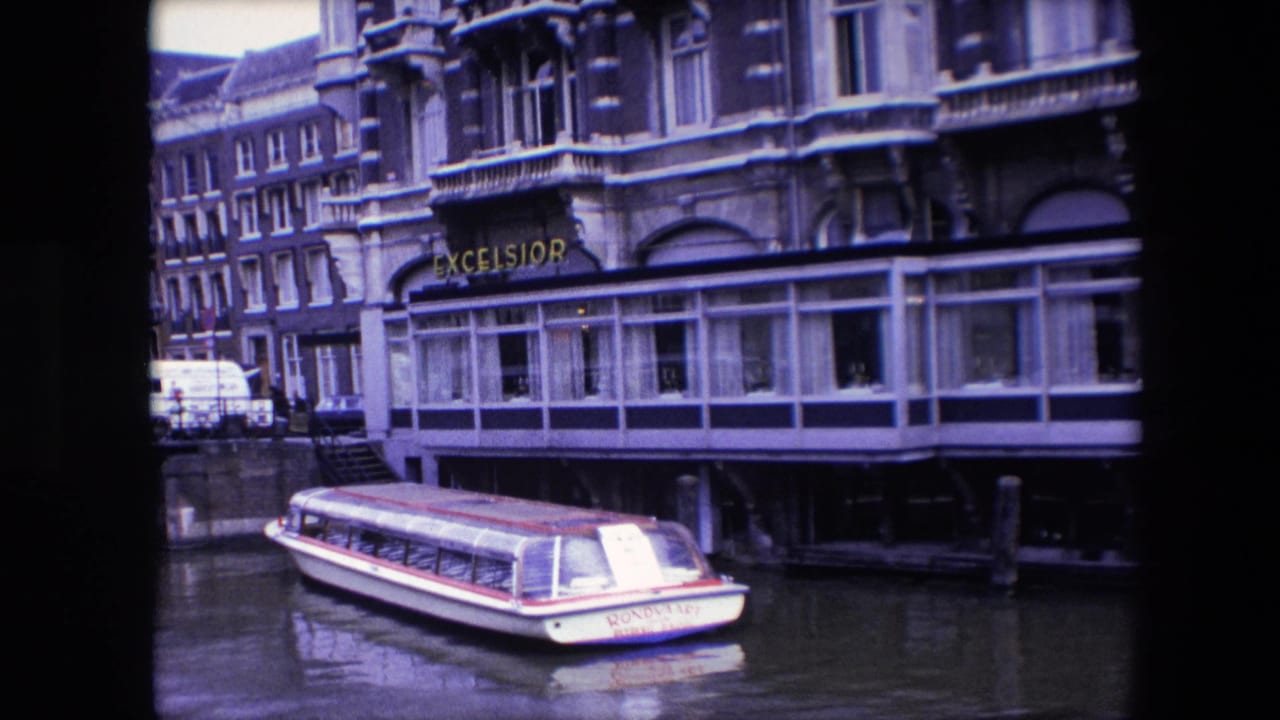 Amsterdam 1967 home-movie frame showing the tour boat closer in the lower foreground, with the Excelsior sign, windowed hotel frontage, and canal houses behind it.