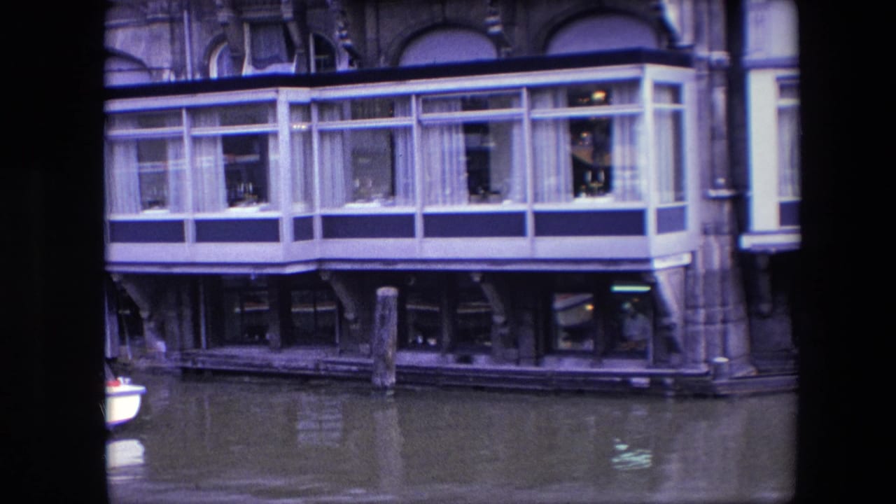 1967 Amsterdam 8mm frame showing a windowed restaurant structure over canal water, with curtained upper windows, pilings, and figures visible inside.