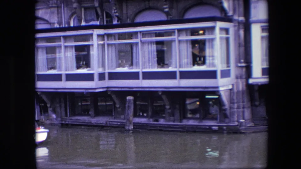 1967 Amsterdam 8mm frame showing a windowed restaurant structure over canal water, with curtained upper windows, pilings, and figures visible inside.