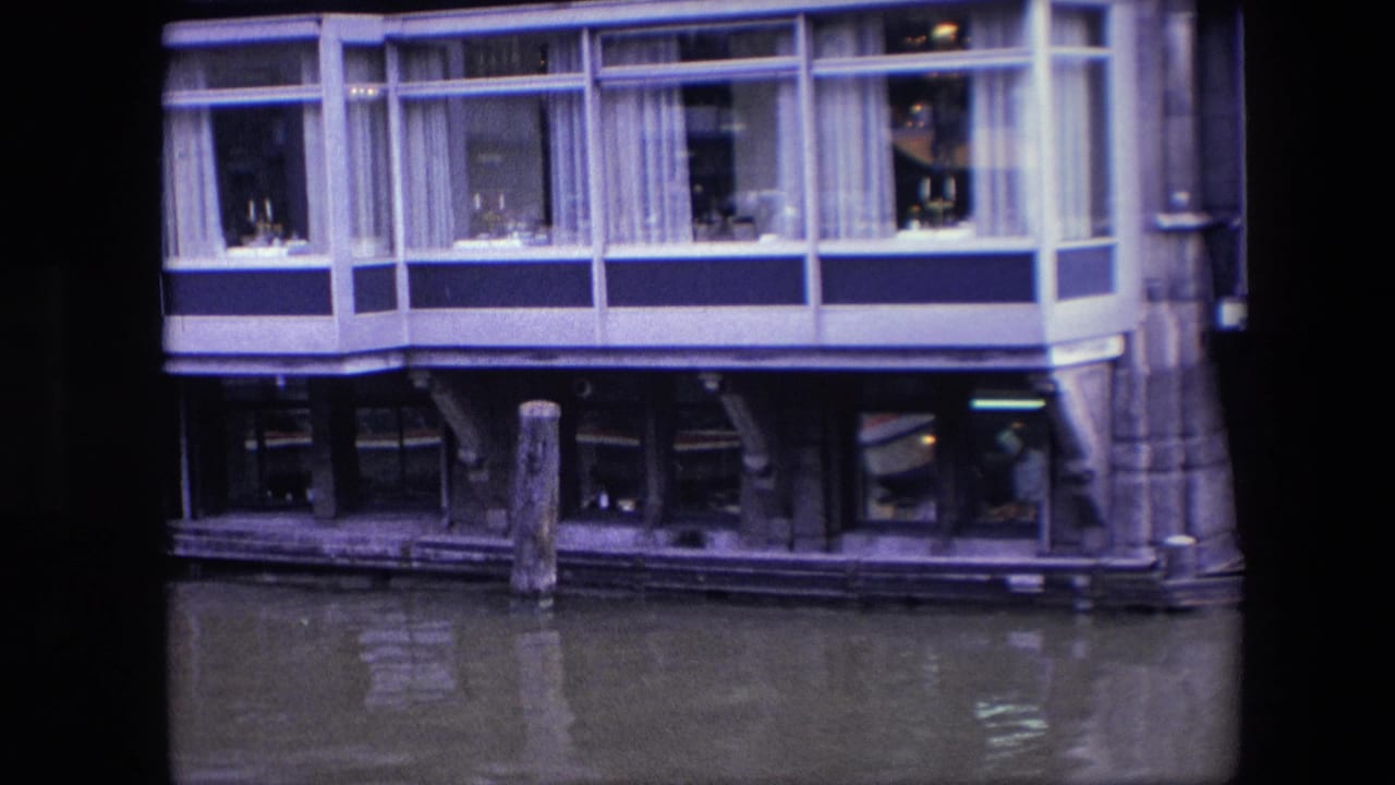 1967 Amsterdam canal view of a restaurant facade built over the water, with upper dining windows, lower interior reflections, pilings, and part of a boat at the left edge.