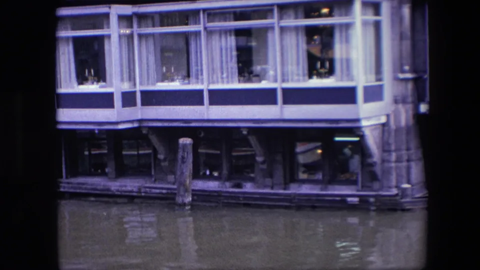 1967 Amsterdam canal view of a restaurant facade built over the water, with upper dining windows, lower interior reflections, pilings, and part of a boat at the left edge.