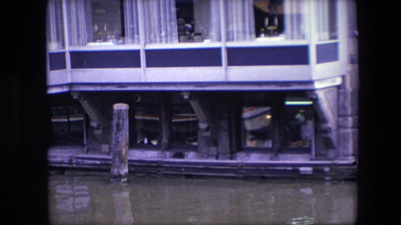 1967 Amsterdam 8mm frame looking toward lower restaurant windows and canal pilings, with interior activity faintly visible behind glass.