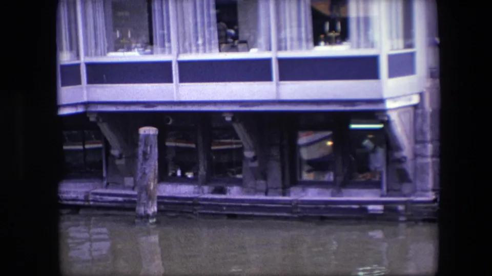 1967 Amsterdam 8mm frame looking toward lower restaurant windows and canal pilings, with interior activity faintly visible behind glass.