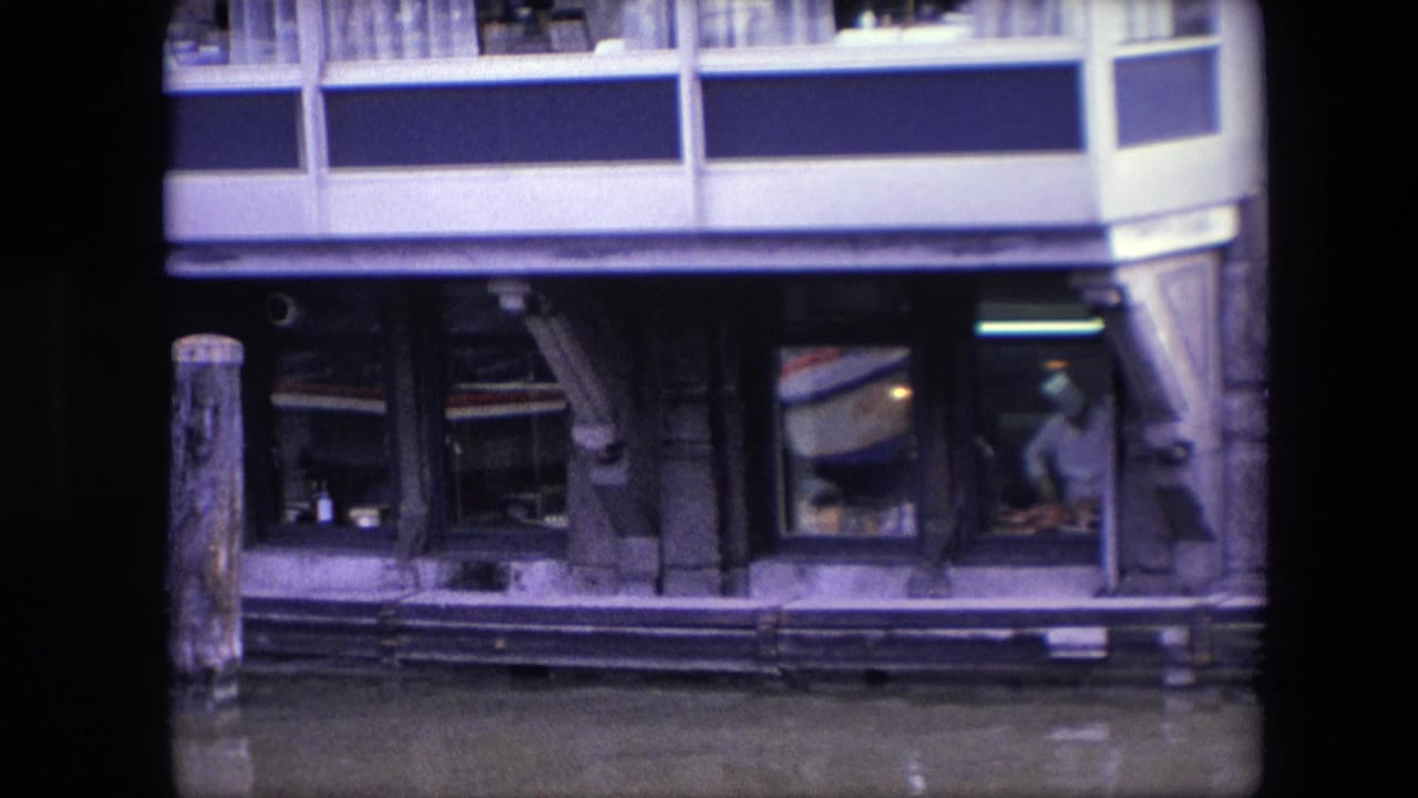 1967 Amsterdam canal restaurant detail showing lower glass windows, a timber piling, water reflections, and a faint interior figure near the right window.