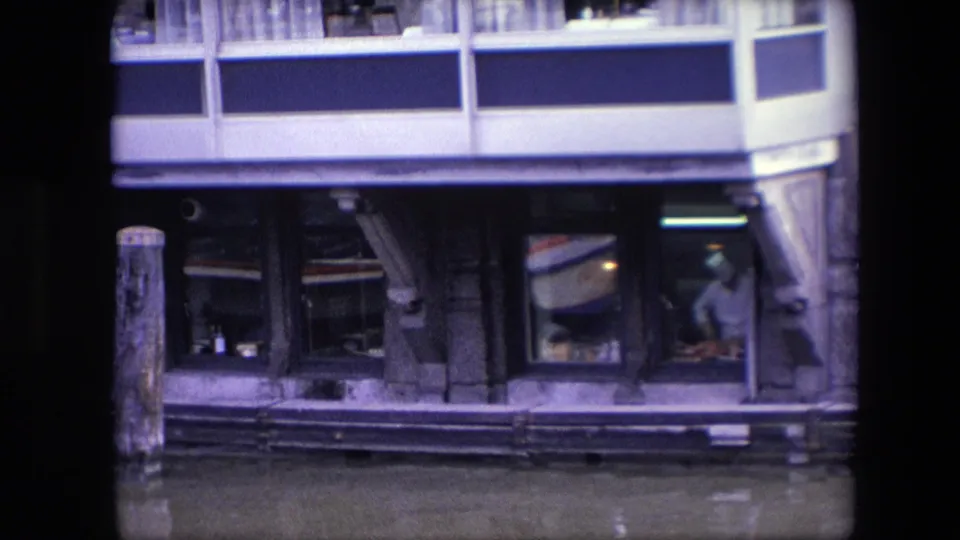 1967 Amsterdam canal restaurant detail showing lower glass windows, a timber piling, water reflections, and a faint interior figure near the right window.