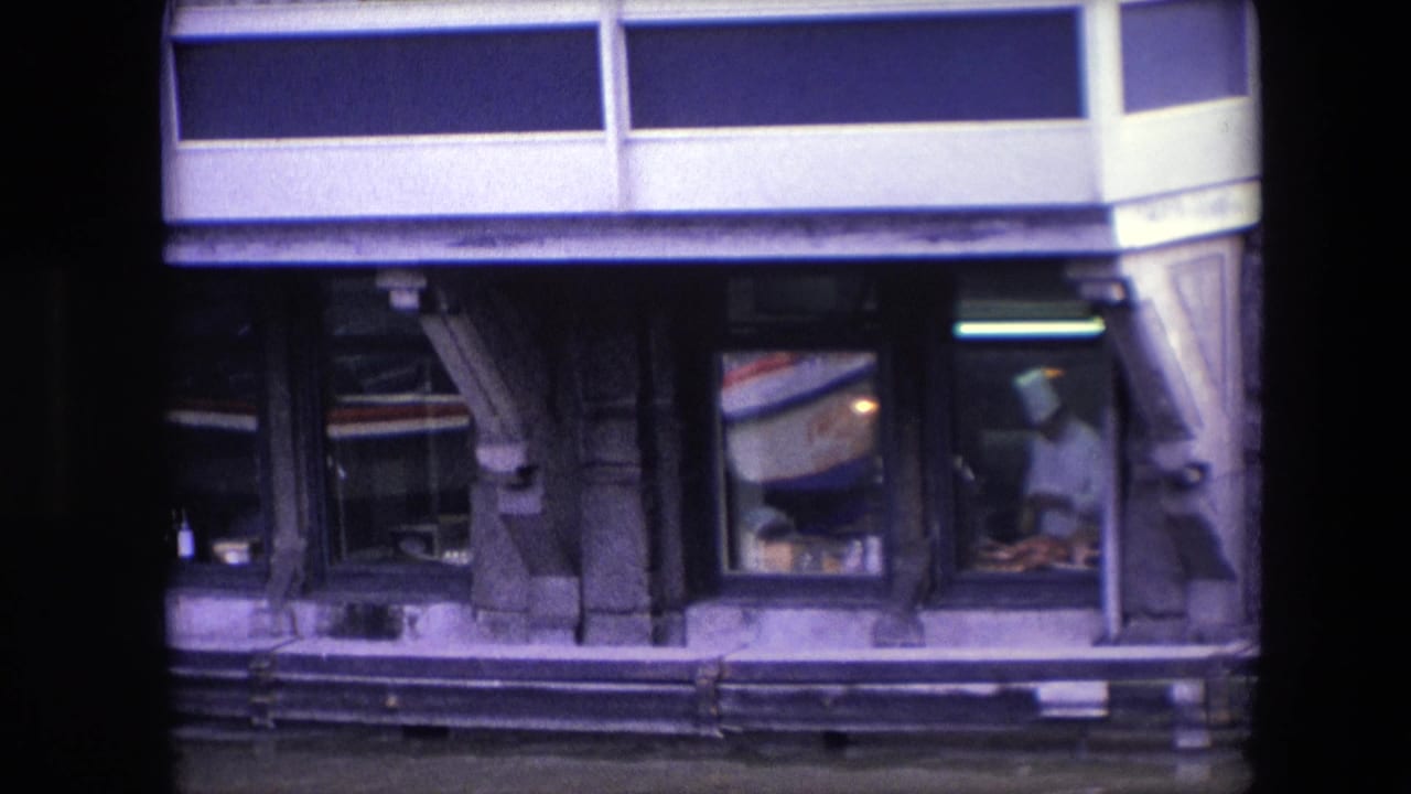 In 1967 Amsterdam 8mm footage, a chef in a white uniform appears through a restaurant window as reflected boat lettering and street-facing facade details cross the glass.