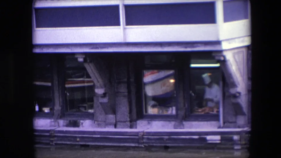 In 1967 Amsterdam 8mm footage, a chef in a white uniform appears through a restaurant window as reflected boat lettering and street-facing facade details cross the glass.