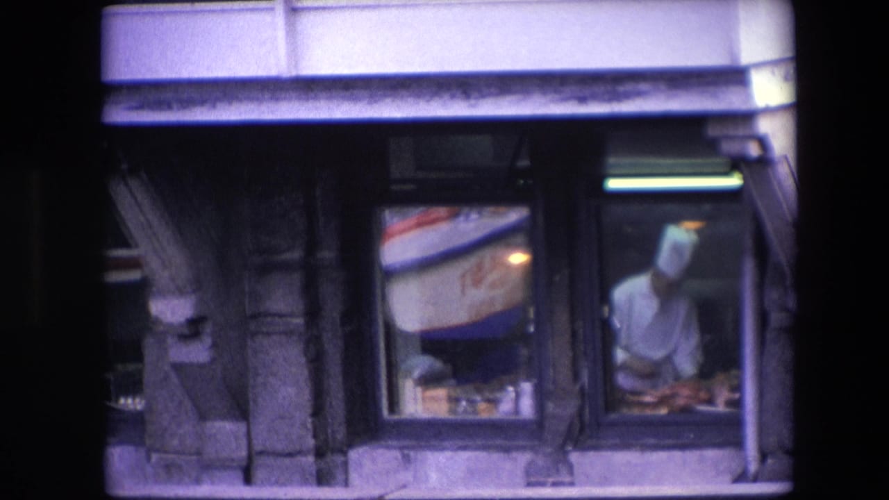 A chef in white kitchen clothing works at a counter inside an Amsterdam restaurant, filmed from outside through dark window frames with reflections in the glass.