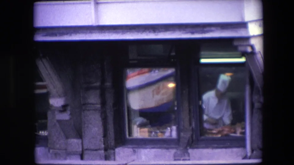 A chef in white kitchen clothing works at a counter inside an Amsterdam restaurant, filmed from outside through dark window frames with reflections in the glass.