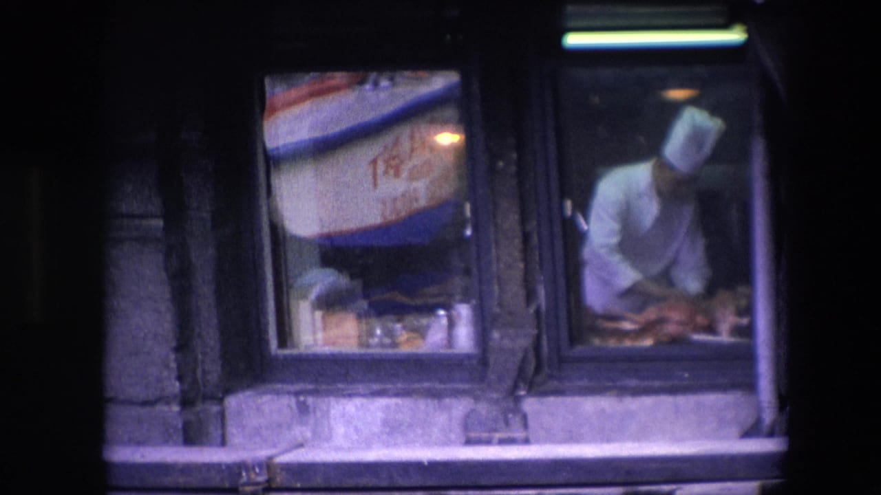 The restaurant window shows reflected boat lettering on the left pane while the chef continues preparing food behind the adjacent glass.