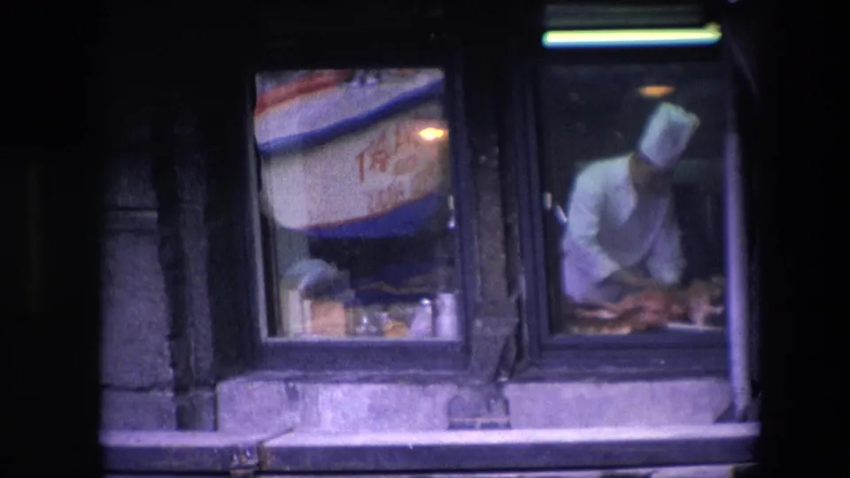 The restaurant window shows reflected boat lettering on the left pane while the chef continues preparing food behind the adjacent glass.
