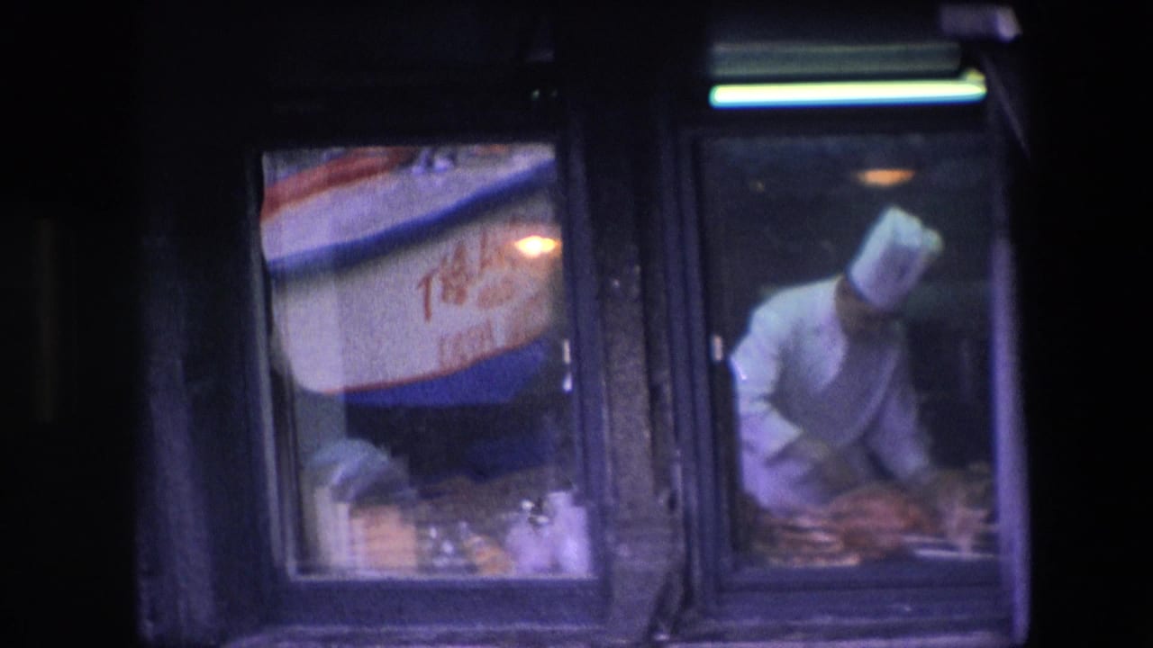 A chef in a tall white hat leans over food preparation inside the restaurant kitchen, seen from the street through reflective glass in the 1967 Amsterdam clip.