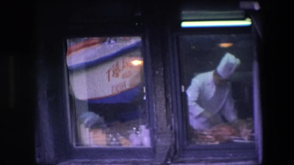 A chef in a tall white hat leans over food preparation inside the restaurant kitchen, seen from the street through reflective glass in the 1967 Amsterdam clip.