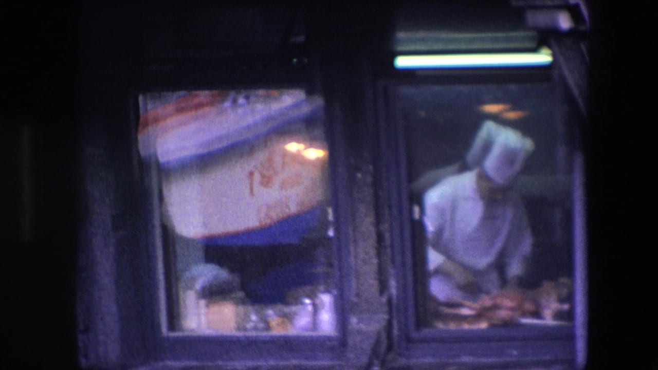 In 1967 Amsterdam footage, a male chef in a white uniform and toque appears behind a restaurant window with food on the counter and a red-and-blue sign nearby.