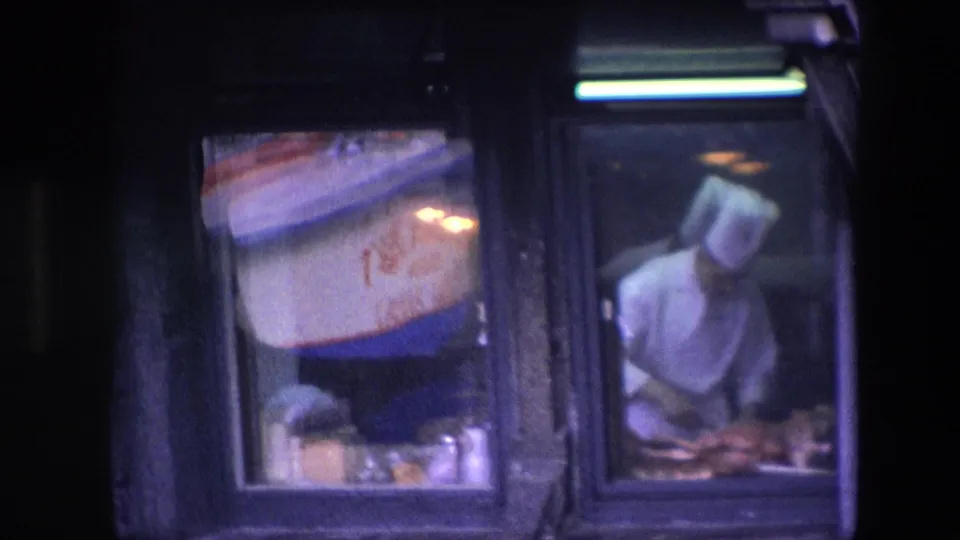 In 1967 Amsterdam footage, a male chef in a white uniform and toque appears behind a restaurant window with food on the counter and a red-and-blue sign nearby.