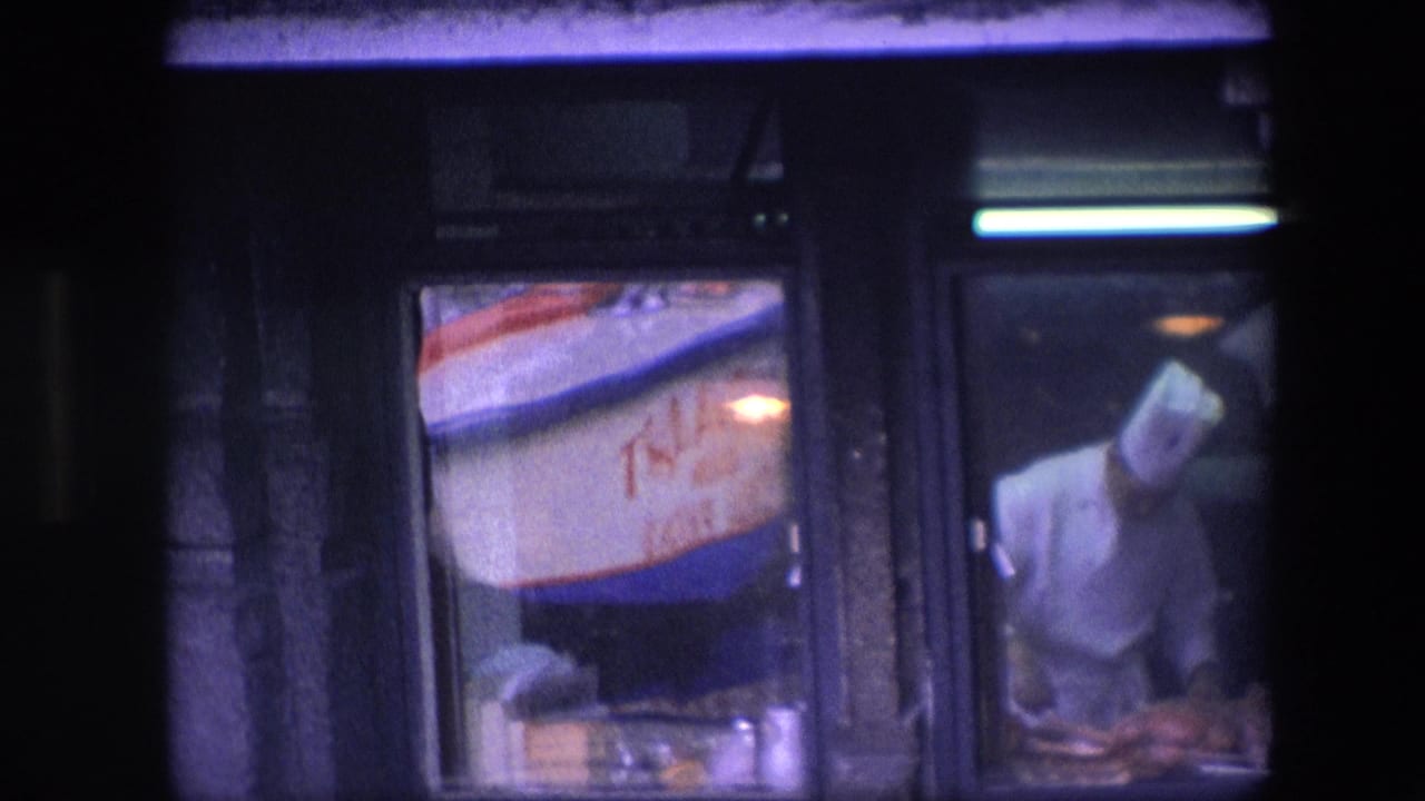 Chef in whites leans over food behind glass beside a lit sign.