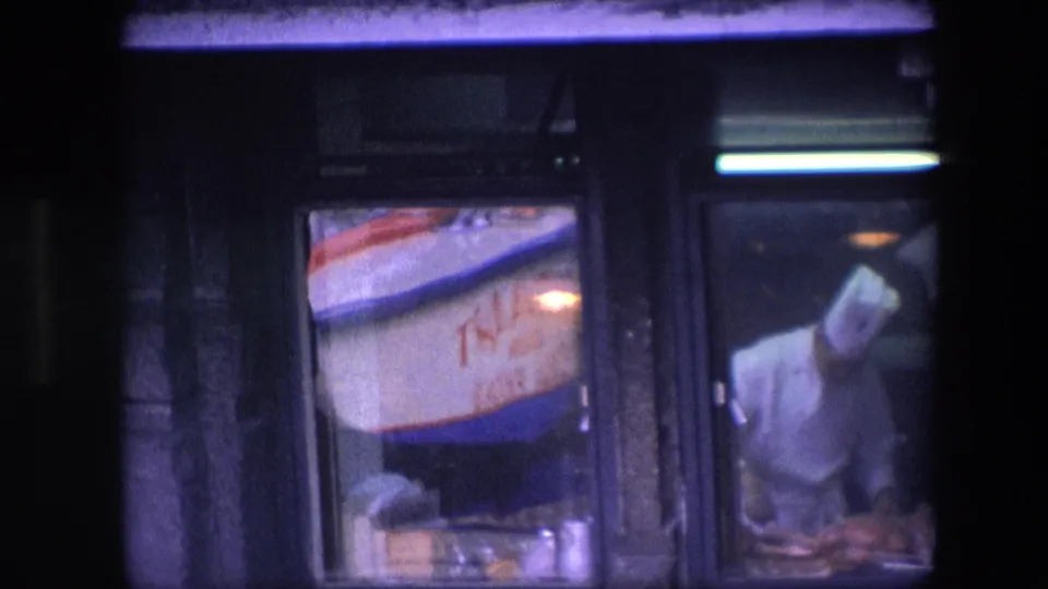 The chef leans forward behind the window, partly obscured by reflections and low light, with a fluorescent tube above and a neighboring red-and-blue sign visible.