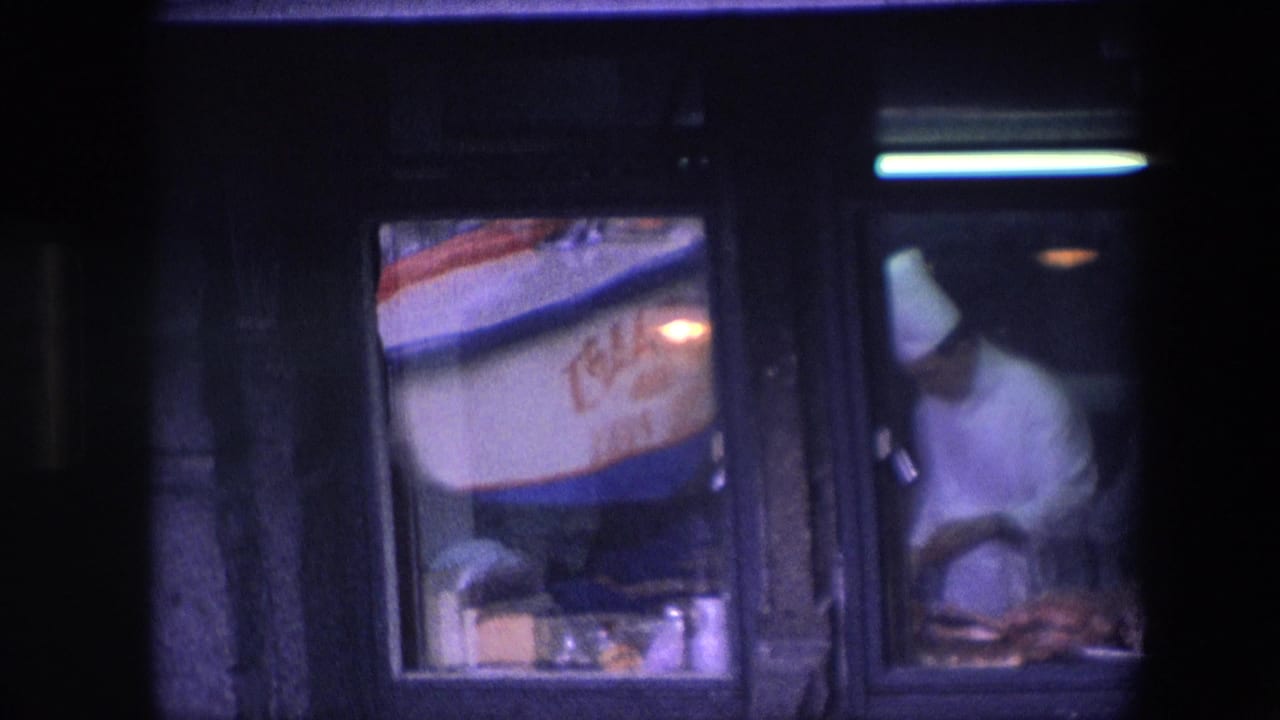 A wider low-light frame shows the restaurant window, the adjacent red-and-blue sign, and the chef in white working at the counter in the background.