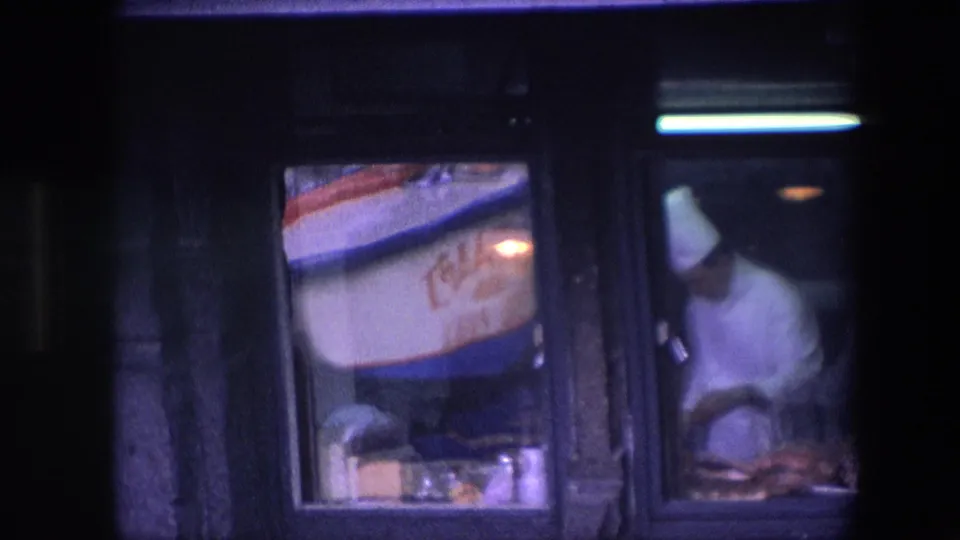 A wider low-light frame shows the restaurant window, the adjacent red-and-blue sign, and the chef in white working at the counter in the background.