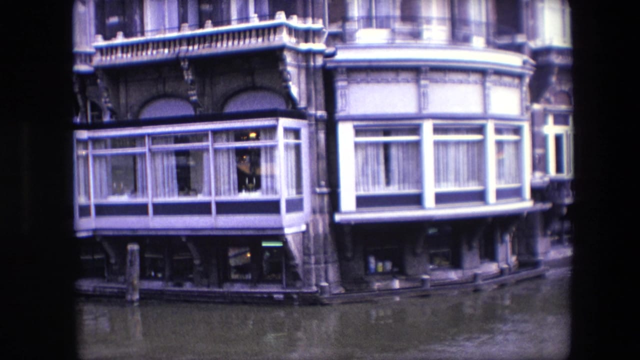1967 Amsterdam canal view showing ornate building details, white-framed bay windows, curtains, lower storefront windows, and muted reflections in the canal.