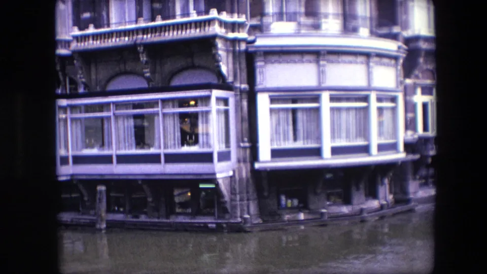 1967 Amsterdam canal view showing ornate building details, white-framed bay windows, curtains, lower storefront windows, and muted reflections in the canal.
